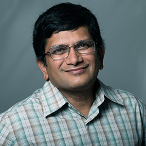 A man in a button up shirt and glasses poses for a photo in front of a cloudy backdrop