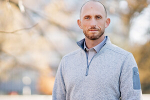 A man wearing a gray sweater poses for the camera while standing outside