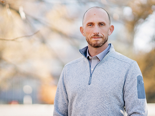 A man wearing a gray sweater poses for the camera while standing outside