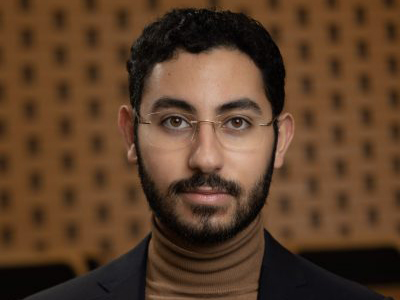 A professional headshot of a young man with a beard and glasses.