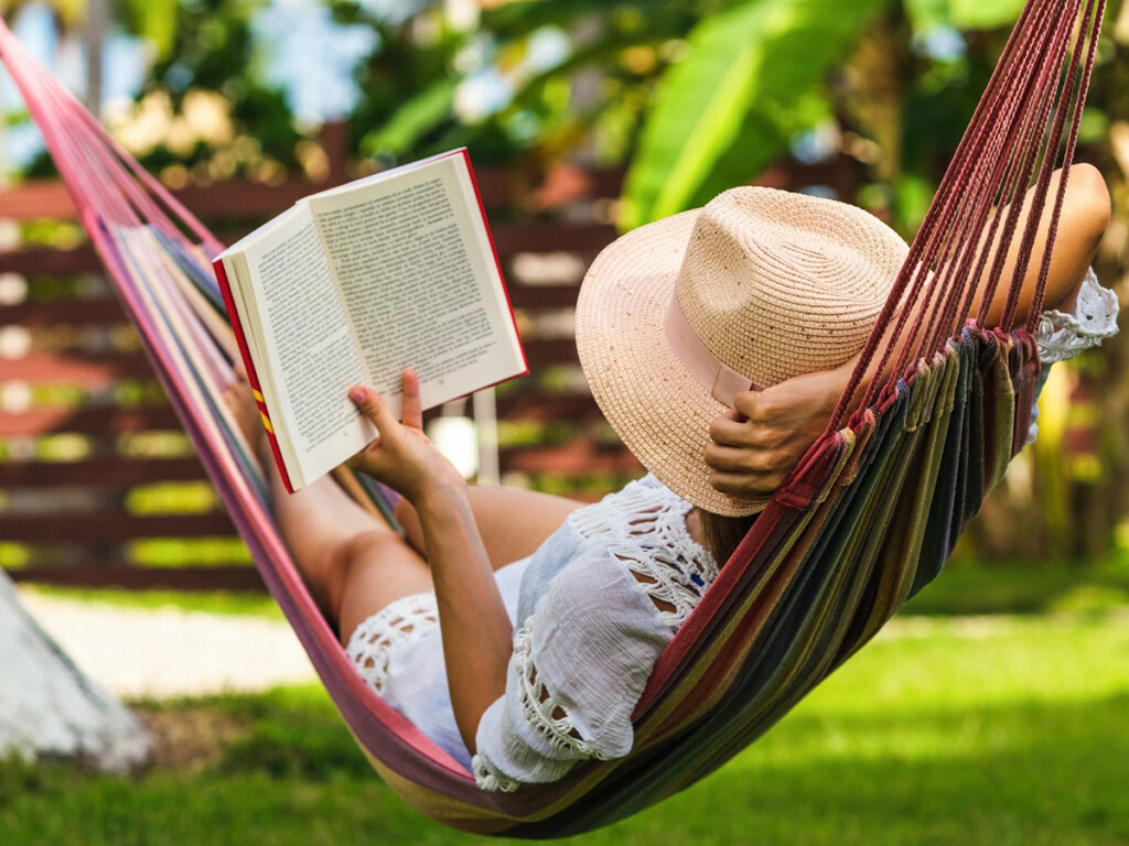A woman wearing a hat is reading a book while laying in a hammock, in background is a beautiful garden and a refreshing sunny day.