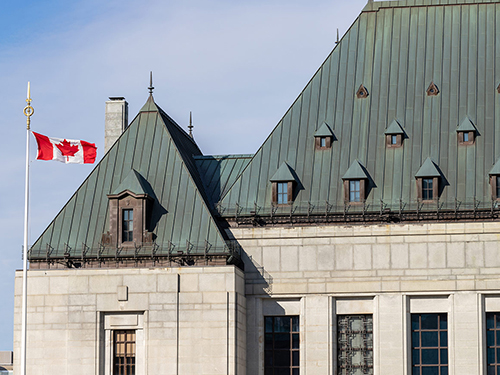 The outside of a large, older building. The Canadian flag can be seen on a large flag pole next to the building.