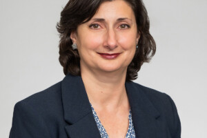 A professional headshot of a woman with dark hair, wearing a dark blazer.