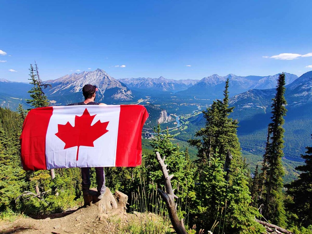 A man holds out a Canadian flag while standing on a mountain top.