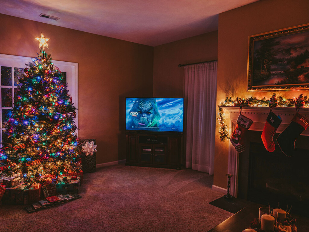 A living room with a Christmas tree, stockings above a fireplace, and a TV with a Christmas movie visible on the screen.