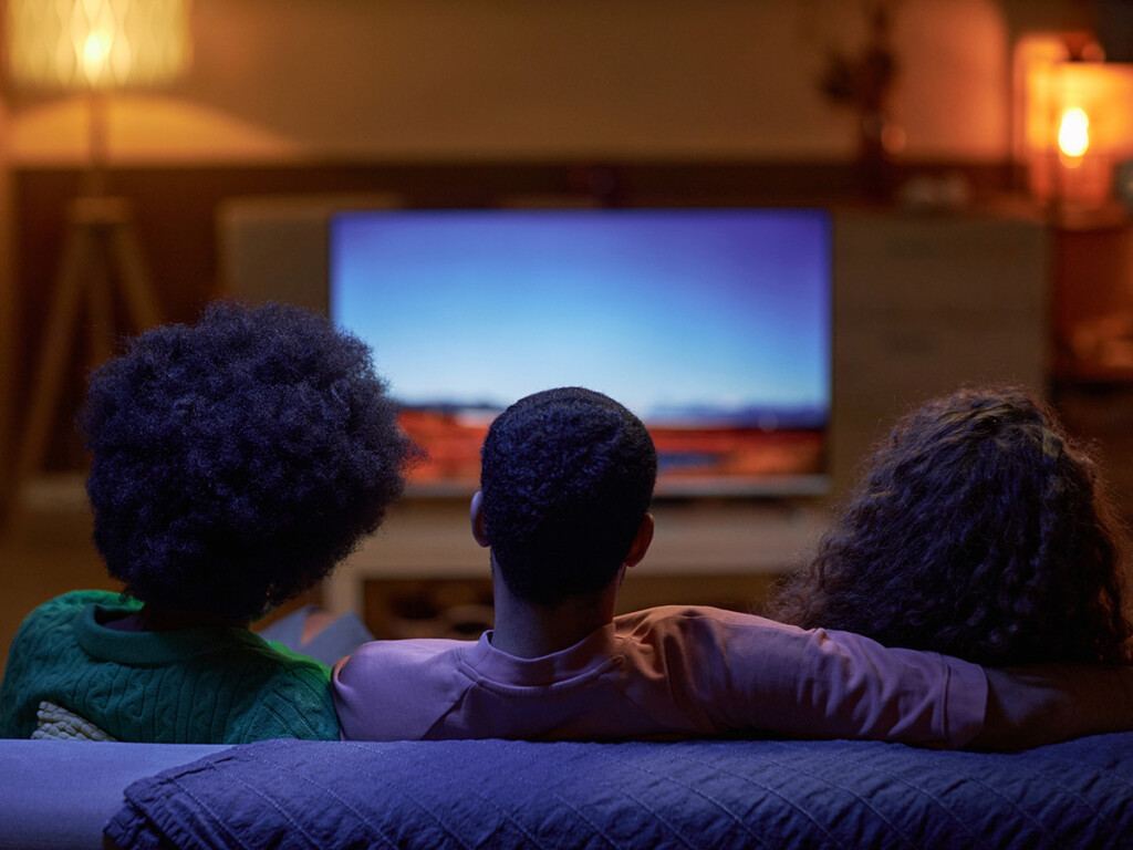 A from behind view of a three people sitting on a couch and watching TV.