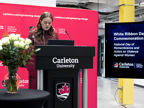 A professionally dressed woman stands at a podium in front of a microphone