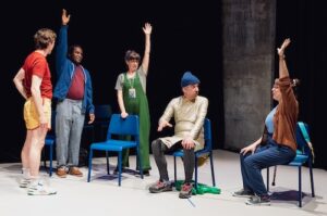 Two people stand onstage, dressed in very colourful clothes, and hold their hands and arms up high. Two sit on chairs, one person staring at the other, who sticks her arm into the air. A fifth person stands with his hands on his hips looking at the rest.