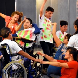 Dancers sit and stand between people in wheelchairs, with everyone moving their arms in all directions.