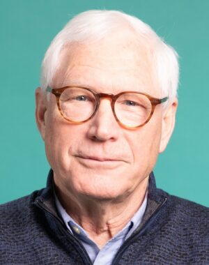 A man who's about 60 years old, wearing with a dress shirt, jacket and glasses, smiles at the camera.