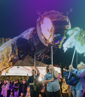 At a street arts festival, a crowd watches four people hold sticks to prop up a Chinese paper dragon that seems to breathe fire.
