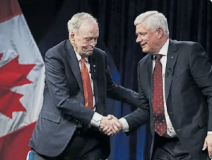 Former prime ministers Jean Chretien and Stephen Harper smile and shake hands, with the Canadian flag behind them.