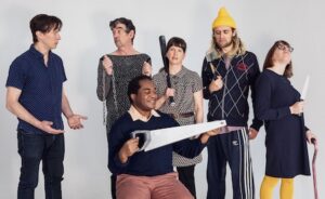 Six actors pose for the camera with various weapons. One holds a saw, another, a bat, and a third, a knife. A fourth holds a chain while looking at a man who's holding his arms out.