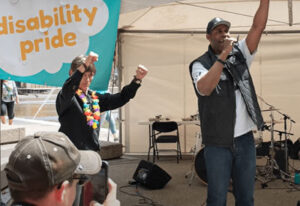 A women with a rainbow wreath around her neck raises her arms in celebration. She stads beside a man with a baseball cap who talks at a microphone.
