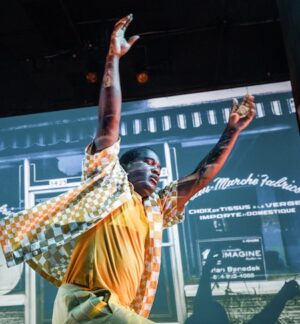 A man walks with his eyes closed and arms stretched above his head. Behind him is a shop window.