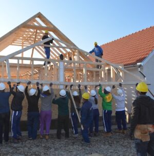 A group of construction workers and volunteers with hard hats build a wooden house.