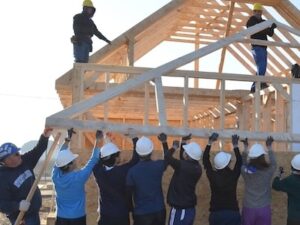 A group of people lift a large, wooden roof onto a wooden house.