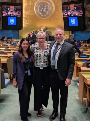 Tara Levandier stands beside two colleagues in a auditorium at the United Nations. Hundreds of desks surround them, each desk labelled with a country's name.