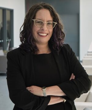 A woman (Jamie), with glasses and long hair, crosses her arms and smiles at the camera.