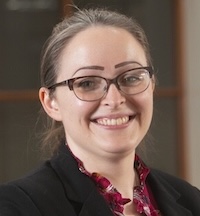 A woman in glasses and brown hair, wearing a read shirt and black blazer, smiles ath the camera.