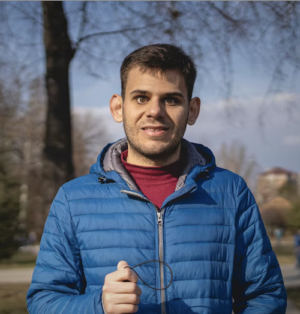 A man with a very short hair and a blue winter jacket smiles at the camera.
