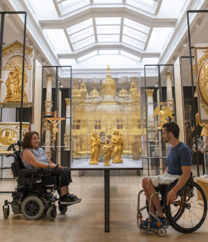 Two people in wheelchairs roll towards each other. There's a massive bright skylight above them, and gold scuptures around them.