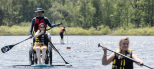A woman in a wheelchair balances on a paddleboard on a lake as she paddles. A man stands behind her paddling on the other side.