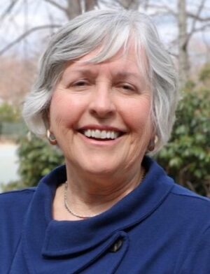 Patricia Bradshaw smiles at the camera. She stands outside in front of green trees, and has grey-white hair and wears a blue sweater and necklace.
