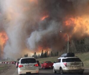 A long line of cars on a road escape a massive wildfire and smoke covering the entire sky.