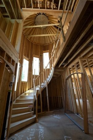 Inside of a partially built wooden house, a beautiful, wooden spiral staircase climbs from the first floor to the second floor surrounded by four bright, wood-framed windows and a domed wooden ceiling.