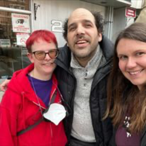 A woman with red hair and a red jacket stands beside a man with a short beard and black jacket and a woman with long brown hair and purple shirt. All three smile at the camera.