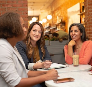 Three people sit at a table at a cafe. Two of them listen intently the third talking.