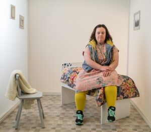 A woman with Down Syndrome wears a pink dress and yellow tights and sits on a tiny bed in a very small room. She sits with one hand over the other on her lap -- and looks at the camera.
