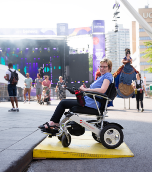 A person in a wheelchair rolls up a yellow ramp to a sidewalk. Behind them is a person walking on stilts.