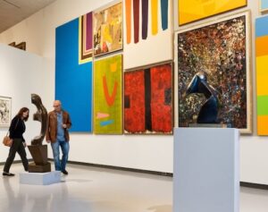 Two people look at a sculpture in an art gallery, with brightly coloured abstract paintings hanging on the wall beside them.