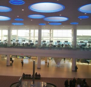 A wide, two-storey airport lobby with huge dome lights on the ceiling people, people sitting on chairs in front of a wall of windows, and more people walking on the lowest floor.