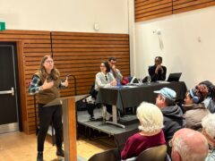Speaker Claire French is seen speaking in front of a podium to event attendees, a panel of other speakers beside her