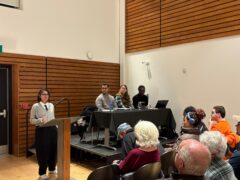 Professor Meena Krishnamurthy is seen speaking in front of a podium to event attendees, a panel of other speakers beside her