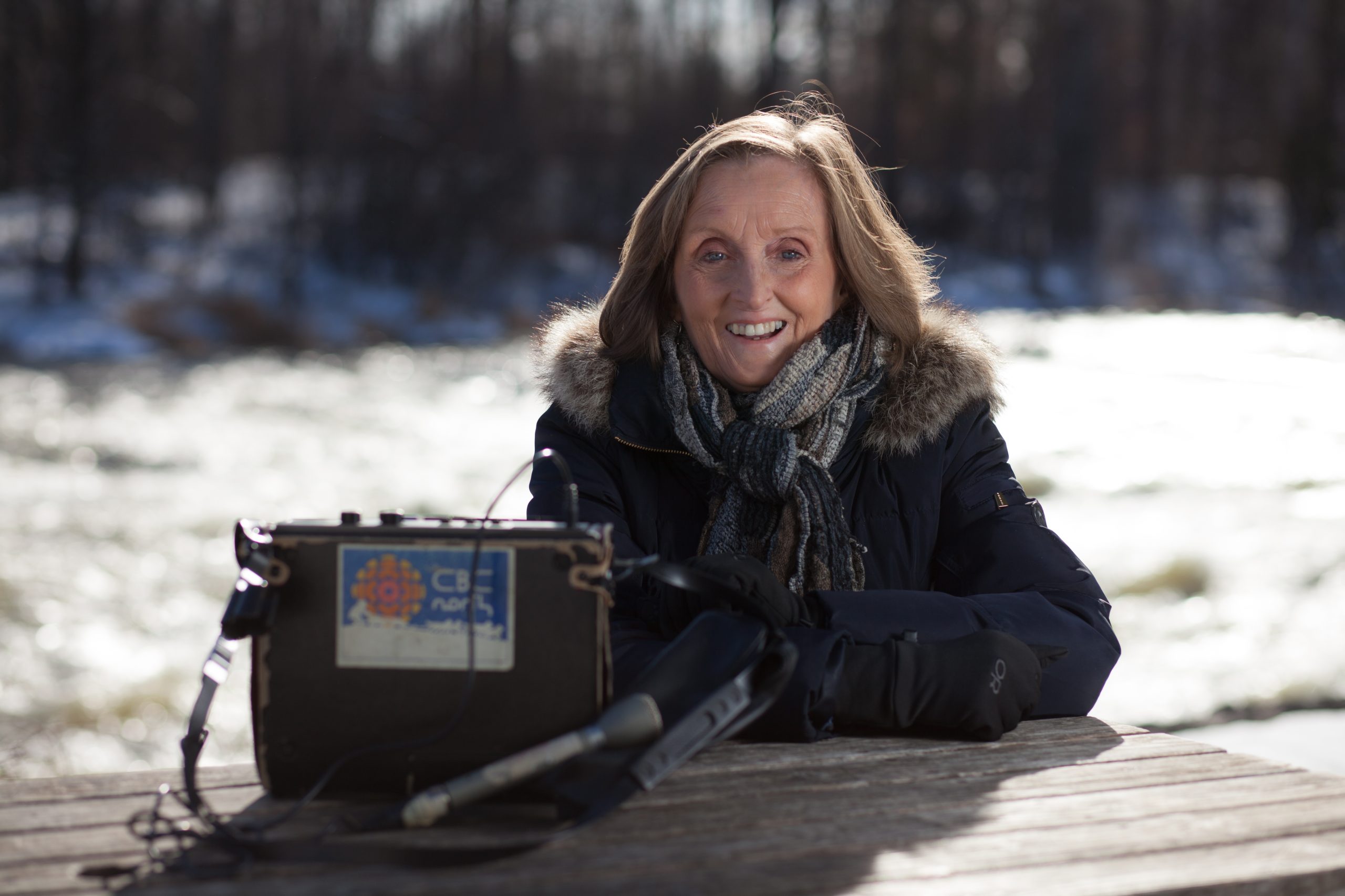 Mary McGuire Sitting Outside At Picnic Table In Winter With CBC Radio Pack On Table