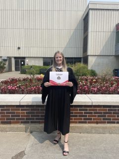 Emily Kelly outdoors, wearing a graduate gown, holding her Carleton Degree and facing the camera.