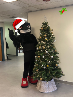 PMC Director Bruce Hamm wearing the Rodney the Raven suit, standing beside a decorated Christmas Tree giving a thumbs up, wearing a santa hat.