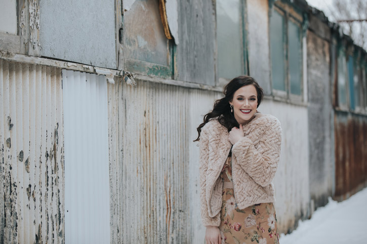 Tessa Virtue pulls a shawl over her shoulders while smiling at the camera next to a barn wall outdoors on a snowy day.