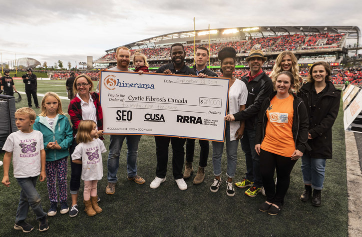 On the field at a Redblacks football game, Carleton students and community members hold a giant cheque for $29,000 made out to Cystic Fibrosis Canada