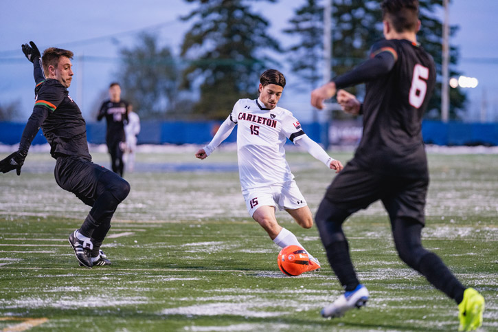 A Ravens men's soccer player, flanked by two opposing players, kicks a ball during an outdoor game in the evening