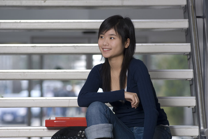 A student of East-Asian descent sits on a set of stairs with textbooks sitting next to her.