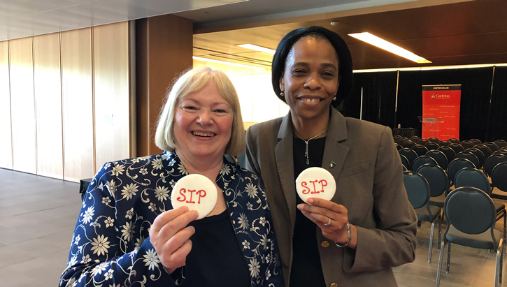 Lorraine Dyke and Patrice Smith stand in front of an empty conference room and hold up cookies with the letters "SIP" written on them.