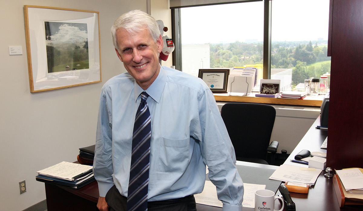 A man wearing a dress shirt and tie leans against a desk while posing for a photo.
