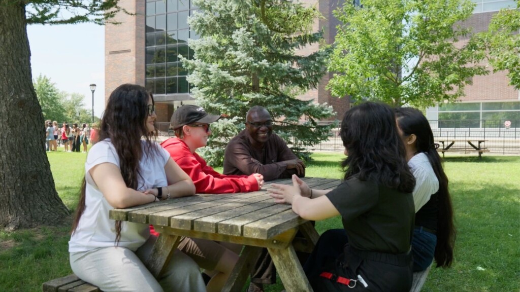 Five people sit around a picnic table on a university campus, talking and smiling outdoors on a sunny day. Trees, grass and a campus building are visible in the background.