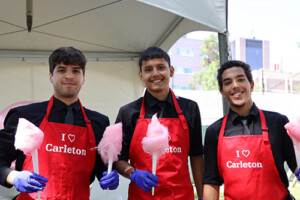 Three people wearing red aprons holding up pink cotton candy in front of a large tent outside.