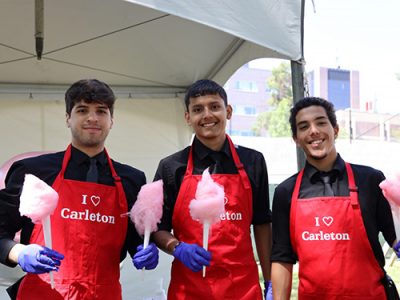 Three people wearing red aprons holding up pink cotton candy in front of a large tent outside.